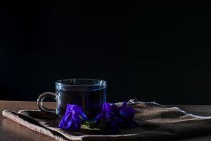Cup of Butterfly pea tea with fresh violet flower on brown tablecloth and wooden table on black background. Healthy beverage for drink. Herbs and medical concept.