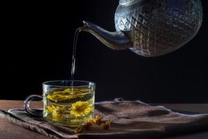 Pour water to a cup of Chrysanthemum tea with dry flower on brown table cloth on black background. Healthy beverage for drink. Herbs and medical concept. photo