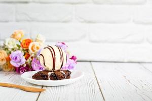 Piece of brownie cake with vanilla ice cream on white small plate on wooden table and with brick wall with copy space.