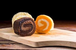 chocolate, vanilla and orange roll cake with whipping cream on  a wooden chopping board and table at the home kitchen with a black background and copy space. Homemade bakery concept. photo
