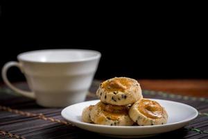 nuez de cajú y galletas de sésamo negro en un plato blanco y una mesa de madera con una taza de café borrosa en el fondo negro. concepto de tiempo de café. foto