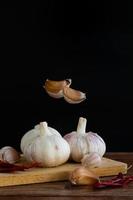 Group of garlic on chopping board and some garlic cloves floating in the air and red dried chilli on wooden table with black background. Copy space for your text. photo