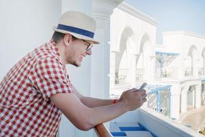 Handsome Caucasian young man standing on balcony of hotel, using mobile phone photo