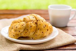 Breakfast concept. Croissants with perilla seeds on white plate and white cup of black coffee on wooden table with green bokeh background. photo