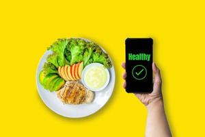 Healthy food concept. Hand holding smartphone with green text healthy with chicken salad and fresh lettuce, avocado, and apple sliced on white plate served with salad dressing on yellow background. photo