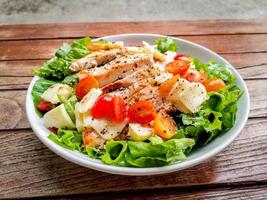 Healthy food concept. Grilled chicken breast salad with fresh red tomato, green lettuce, and mozzarella cheese on a white dish on wooden table. photo