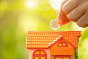 Hand put the coin to piggy bank Home Model on green nature blur background. Money savings concept photo