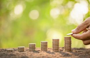 Hand putting coins in stack on wooden plank with green blur background. Money, Finance or Savings concept photo