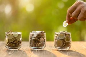 Hand putting coins into clear money jar on wooden table with green blur light background. Savings concept photo