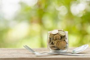 Coins in clear money jar, fork and spoon on wooden table with green blur light background. Savings money for eating concept photo