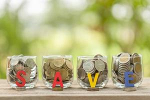 Hand putting coins into clear money jar on wooden table with green blur light background. Savings concept photo