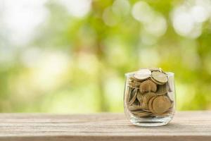 Hand putting coins into clear money jar on wooden table with green blur light background. Savings concept photo