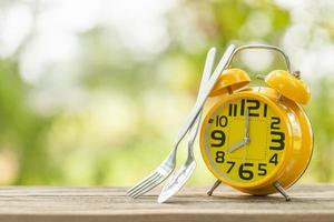 reloj despertador amarillo, tenedor y cuchara sobre una mesa de madera con fondo verde borroso de la naturaleza al aire libre. ocho en punto, hora de comer concepto foto