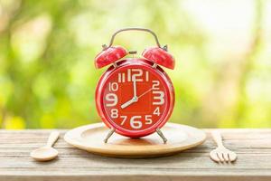 reloj despertador rojo, tenedor y cuchara sobre una mesa de madera con fondo verde borroso de la naturaleza al aire libre. ocho en punto, hora de comer concepto foto