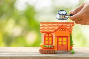 Hand holding blue stethoscope and orange house model on wooden table. Home and building checking photo