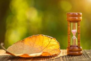 reloj de arena vintage o reloj de arena con hoja de árbol seca sobre una mesa de madera, fondo de desenfoque de naturaleza verde foto