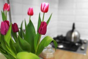 Tulips bouquet close up at home standing on a wooden countertop in the kitchen. Modern white u-shaped kitchen in scandinavian style. Open shelves in the kitchen with plants and jars. photo