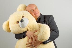 An adult bald man in a black suit holds a large toy bear on a gray background photo