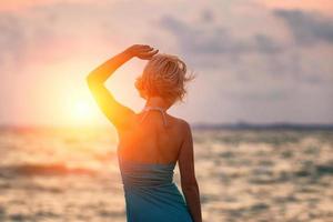 A young slender girl stands on the beach at sunset in a blue long dress, a beautiful figure photo