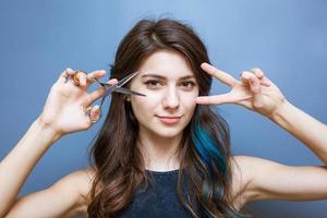 Cute young woman with brunette with blue strands of hair holding scissors near photo