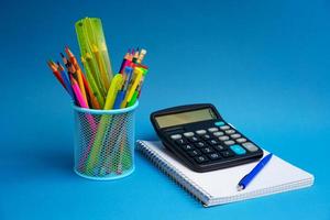 holder with pens and pencils and  notebook with calculator on  blue background. photo