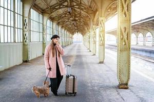 Travel concept. At station, young tourist with dog goes and drags suitcase photo