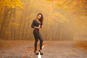 bella joven en el bosque de otoño en la carretera posando con chaqueta negra y polainas foto