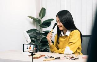 Beautiful asian woman blogger using camera phone recording vlog video live streaming and showing how to make up cosmetics at home. photo