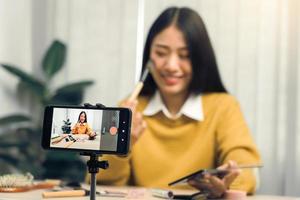 Beautiful asian woman blogger using camera phone recording vlog video live streaming and showing how to make up cosmetics at home. photo