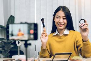 Beautiful asian woman blogger using camera phone recording vlog video live streaming and showing how to make up cosmetics at home. photo