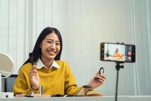 Beautiful asian woman blogger using camera phone recording vlog video live streaming and showing how to make up cosmetics at home. photo