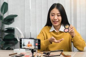 Beautiful asian woman blogger using camera phone recording vlog video live streaming and showing how to make up cosmetics at home. photo