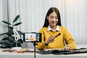 Beautiful asian woman blogger using camera phone recording vlog video live streaming and showing how to make up cosmetics at home. photo
