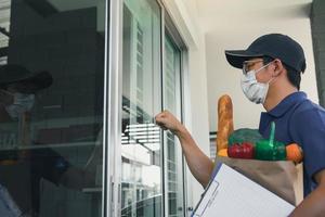 hombre asiático con una máscara mientras sostiene una bolsa de comida llamando a la puerta principal. foto