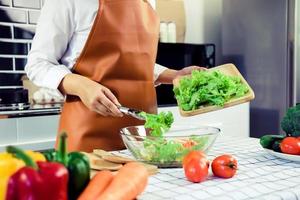 ama de casa asiática está usando pinzas para llevar la ensalada en la tabla de cortar de madera a la taza de ensalada en la cocina. foto