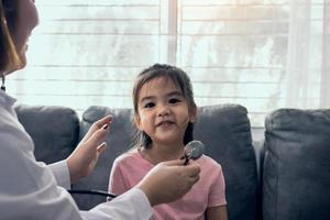 Asian girl child is smiling up and sits on sofa at home during a medical examination while female medical professional is using a stethoscope to listen to her heartbeat. photo