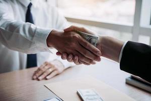 Two corporate businessmen shaking hands while one man places money on document in office room with corruption concept. photo