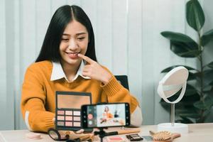 Beautiful asian woman blogger using camera phone recording vlog video live streaming and showing how to make up cosmetics at home. photo