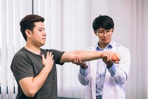 Asian young male physiotherapist helping patient with lifting dumbbells exercises in clinic. photo