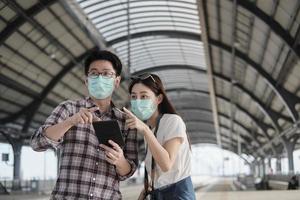 Couple Asian tourists with face masks search information, find travel locations by mobile phone map at a train station in Thailand, passenger trip lifestyle, casual transportation, journey vacation. photo
