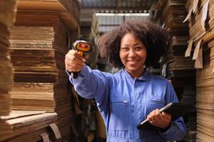 One African American female worker in safety uniform using bar code scanner in front of a camera, check shipment orders at parcels warehouse, cardboard manufacture factory for the packing industry. photo