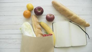 Full paper bag of food products with blank little notebook on wooden table, top view photo