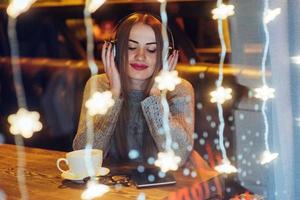 Young beautiful woman sitting in cafe, drinking coffee. Model listening to music. Christmas, new year, Valentines day, winter holidays concept photo