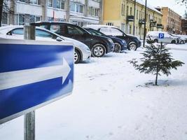 flecha blanca en la señal de carretera azul que apunta a un pequeño pino en el estacionamiento cubierto de nieve foto