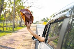 feliz disfrute y libertad en el viaje de viaje con la mano levantada y sosteniendo un sombrero de vaquero fuera del auto de la ventana en las vacaciones de verano foto