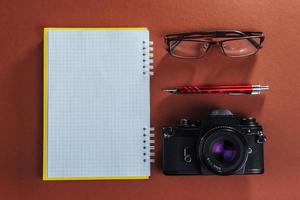 camera, glasses and notepad and pencil on a brown wooden background