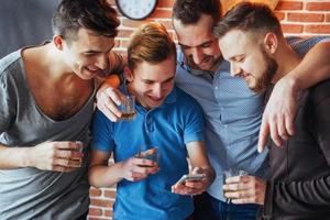 viejos amigos alegres se comunican entre sí y miran por teléfono, vasos de whisky en el pub. concepto de estilo de vida de entretenimiento. personas conectadas wifi en una reunión de mesa de bar foto