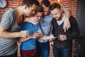 viejos amigos alegres se comunican entre sí y miran por teléfono, vasos de whisky en el pub. concepto de estilo de vida de entretenimiento. personas conectadas wifi en una reunión de mesa de bar foto