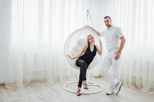 Young couple at white room. Girl sits in a hanging chair photo