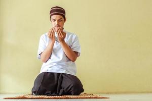 Front view of young asian muslim praying to allah. Islam guy sitting with begging hand gesture on the prayer mat with copy space photo